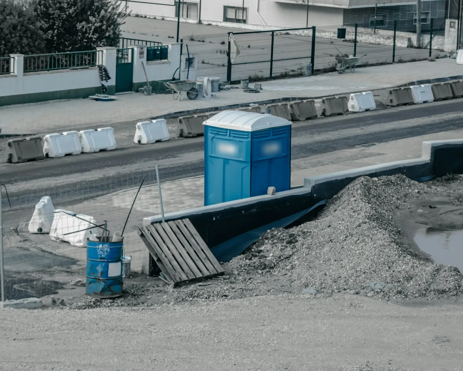 A portable toilet on an active jobsite, beside traffic barriers and a pile of gravel